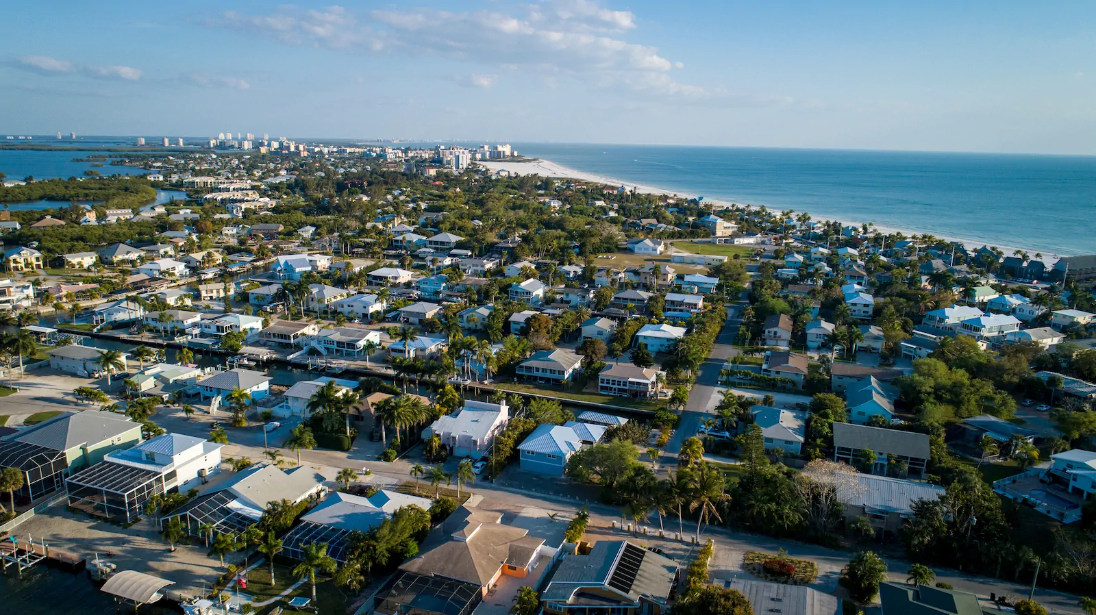 An aerial sweep of a coastal Lee County neighborhood, canal-front homes and screened pools giving way to the white sand and the open Gulf.